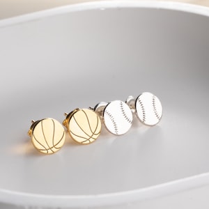 May include: A collection of sports-themed stud earrings. Two pairs of earrings are visible: gold basketball earrings and silver baseball earrings. The basketball earrings have a gold finish with black lines. The baseball earrings are white with red stitching.
