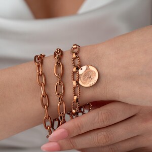 May include: Copper-colored bracelets with varying chain link designs. One bracelet features a round charm with a detailed cat face engraving. The bracelets are worn on a wrist, showcasing the warm tones of the metal.