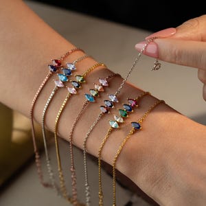 Könnte beinhalten: Eine Sammlung feiner Armbänder, jedes mit einer Kette und einer Reihe farbiger, marquiseförmiger Edelsteine. Die Armbänder sind in verschiedenen Metalltönen gehalten, darunter Gold, Silber und Roségold, mit Edelsteinen in Rot, Blau, Grün und Rosa.