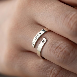 May include: A silver wrap-around ring, engraved with the name "Sophia" on one band and a small heart on the other. The ring's design allows it to wrap around the finger. The ring is shown on a person's finger, showcasing its modern, personalised style.