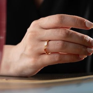 May include: A gold-colored ring with two rectangular gemstones, one white and one red. The ring is worn on a finger, with the hand in the foreground and a dark background.