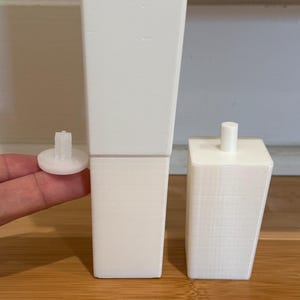 May include: White furniture legs and a small white connector. The legs are rectangular with a slight taper. One leg is shown with a connector attached. The other leg is shown without a connector. The legs are on a wooden surface.