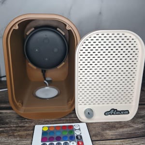 Peut inclure: Un boîtier de haut-parleur marron avec un haut-parleur intelligent noir à l'intérieur, à côté d'un couvercle de haut-parleur blanc avec le mot "Alexa" imprimé dessus. Une télécommande avec des options de couleurs est devant.