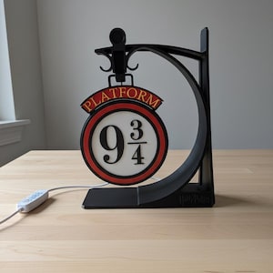 Peut inclure: Un panneau noir et rouge de la plateforme 9 3/4 avec un design circulaire et le texte "PLATFORM" en rouge. Le panneau est soutenu par un support incurvé noir. Un cordon blanc avec un boîtier de commande rectangulaire est attaché au panneau.