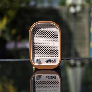May include: A retro-style speaker with a brown frame and a white perforated front. The speaker has a volume knob and the word "Siri" printed on the front. The speaker is reflected on a glossy surface.