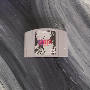 May include: A gray Nintendo 64 game cartridge featuring the game "The Legend of Zelda: Ocarina of Time." The label displays the game's title in English and Japanese, with a graphic of a sword-wielding figure.