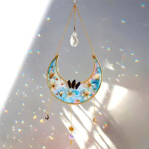 Può includere: Un acchiappasole a forma di mezzaluna con cornice e catena dorate. La luna è riempita di resina blu, fiori bianchi e cristalli scuri. Un cristallo trasparente e ciondoli pendenti completano il design. L'acchiappasole è progettato per essere appeso.