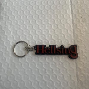 May include: A keychain featuring the word "Hellsing" in red lettering against a black base. It has a silver metal ring and chain. The background is a white bubble wrap envelope.