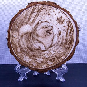 Könnte beinhalten: Ein Holzbrandkunstwerk auf einer kreisförmigen Holzscheibe. Das Kunstwerk zeigt einen Zwergspitzhund in einer Waldszene, der nach einem fallenden Ahornblatt greift. Das Stück wird auf einem transparenten Kunststoffständer ausgestellt.