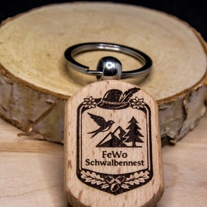 Könnte beinhalten: Ein Holzschlüsselanhänger mit einem silbernen Schlüsselring. Der Holzanhänger zeigt ein graviertes Design eines Hutes, Vogels, Bergen und Bäumen mit dem Text "FeWo Schwalbennest". Der Schlüsselanhänger liegt auf einem Holzscheiben-Hintergrund.