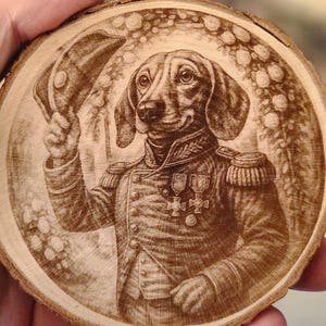 Könnte beinhalten: Eine Holzscheibe mit einer detaillierten Gravur eines Dackels in Militäruniform, der einen Hut hält. Der Hund ist von einem floralen Rand umgeben, mit komplizierten Details in der Holzmaserung.