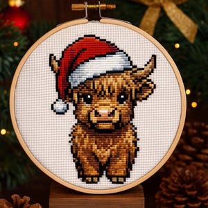 May include: A cross-stitch of a Highland cow wearing a Santa hat. The cow is brown with black eyes and a pink nose. The Santa hat is red and white. The cross-stitch is in a wooden embroidery hoop, with a wooden stand.