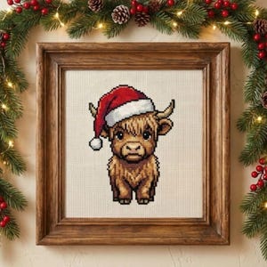 May include: A framed cross-stitch of a Highland cow wearing a Santa hat. The artwork is in a wooden frame and is surrounded by a festive garland of greenery, red berries, and fairy lights. A charming holiday decoration.