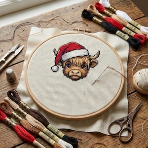 May include: A cross-stitch of a Highland cow wearing a Santa hat, displayed in a wooden hoop. The image shows embroidery floss in various colours, scissors, and a needle, indicating a crafting project.