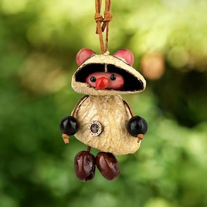 Puede incluir: Adorno artesanal con forma de criatura con cuerpo beige, brazos de cuentas negras y pies de semillas. Tiene una nariz roja, orejas rosas y un cordón de cuero marrón para colgar. El fondo es verde suave.