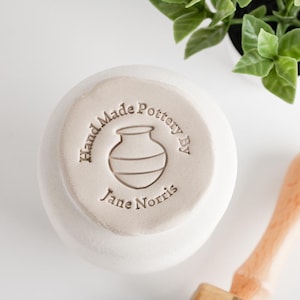 May include: A handmade, off-white pottery bowl with the text "Hand Made Pottery By Jane Norris" stamped on the bottom. The stamp includes a simple illustration of a vase. The bowl is set against a white background.