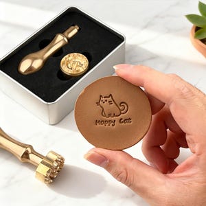 May include: A gold-colored wax seal stamp set. The set includes a stamp handle, a round stamp with a cat design and the text "Happy Cat", and a metal stamp tool. The stamp is shown on a brown leather circle.