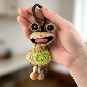 Puede incluir: Un llavero caprichoso con forma de rana hecho de materiales naturales. La rana tiene ojos grandes y oscuros, una boca sonriente con una lengua rosa y una pequeña bolsa verde de ganchillo. El llavero tiene un cordón marrón para colgar.