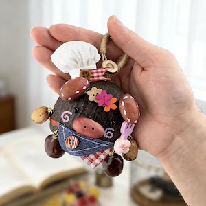 Puede incluir: Un llavero artesanal y caprichoso con un diseño temático de chef. El llavero tiene una base marrón oscuro con elementos decorativos adjuntos, que incluyen un gorro de chef blanco, un babero de mezclilla y varios amuletos con forma de comida. El llavero está sujeto a un lazo de fibra natural.