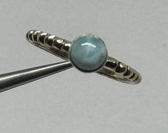 May include: A silver ring with a light blue, round gemstone. The band has a textured design. The ring is held by a pair of tweezers against a white background. The gemstone is approximately 0.25 inches in diameter.
