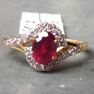 Könnte beinhalten: Ein goldener Ring mit einem großen ovalen Rubin in der Mitte, umgeben von einem Kranz aus kleineren Diamanten. Das Ringband ist geteilt und mit zusätzlichen Diamanten verziert, was einen funkelnden, eleganten Look erzeugt.