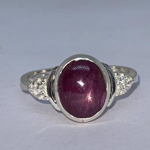 May include: A silver ring featuring a large, oval, deep red gemstone. The gemstone has a star-like effect. Small, clear stones are set on either side of the main stone, adding sparkle to the ring. A classic piece of jewelry.