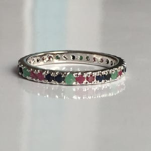 Peut inclure: Une bague en argent ornée d'une rangée continue de pierres précieuses alternées rouges, vertes et bleues. La bague est posée sur une surface blanche, les pierres étant espacées uniformément.