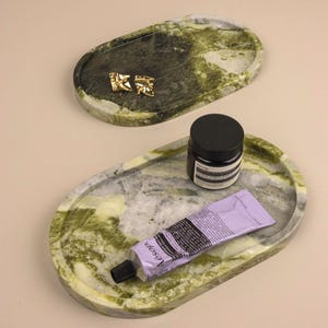 May include: Two oval-shaped green marble trays with natural veining. The top tray holds two gold-toned geometric earrings. The bottom tray holds a black jar with a black lid and a lavender-colored tube of Aesop hand cream with black text.