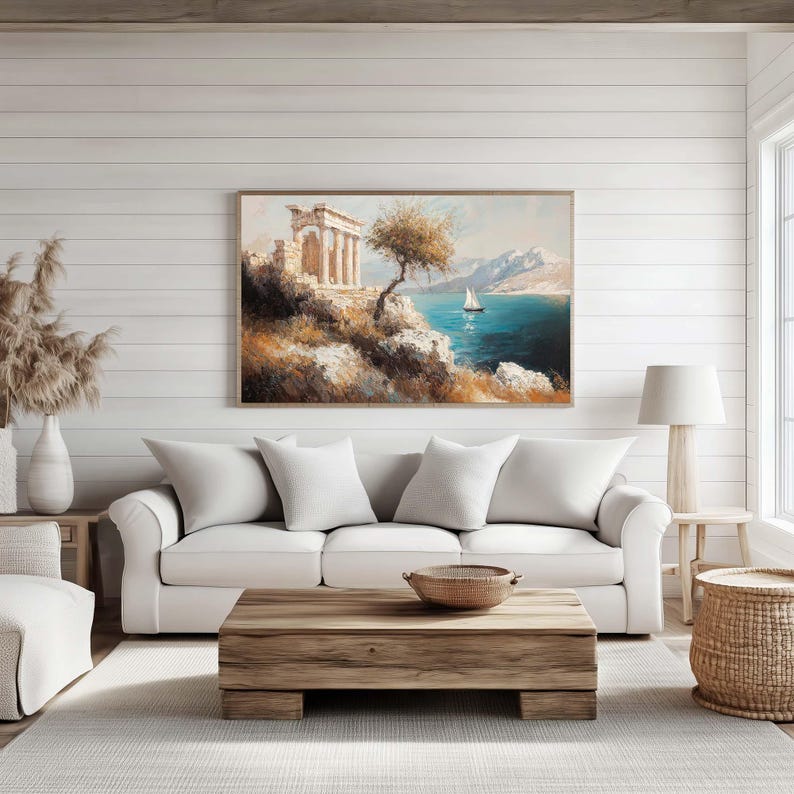 Puede incluir: Una pintura de paisaje enmarcada representa una ruina cl&aacute;sica en un acantilado con vistas a un mar turquesa con un velero. La obra de arte cuelga sobre un sof&aacute; blanco con cojines a juego. Una mesa de centro de madera y una cesta de mimbre completan la decoraci&oacute;n.