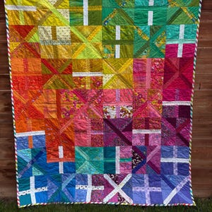 May include: A rectangular patchwork quilt featuring a vibrant rainbow gradient of colors. The design consists of square blocks, each containing a white cross pattern. The quilt is finished with a multi-colored striped binding along the edges.