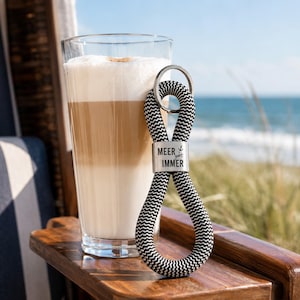 Könnte beinhalten: Ein schwarz-weiß gemusterter Schlüsselanhänger aus Seil mit einem silbernen Metallverschluss, der mit dem Text MEER geht IMMER und einem kleinen Anker-Symbol graviert ist, neben einem hohen Glas mit einem geschichteten Kaffeegetränk.