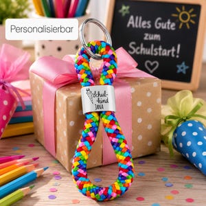 Può includere: Un portachiavi con un cordino intrecciato nei colori dell'arcobaleno, tra cui rosso, arancione, giallo, verde, blu e viola. Una perlina rettangolare in metallo argentato è centrata sul cordino, incisa con un disegno di un cono e il testo Schulkind JANA.