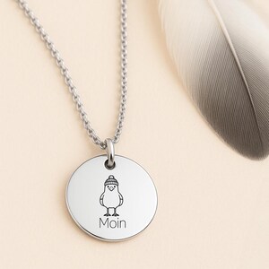 Könnte beinhalten: Eine silberfarbene Halskette mit Anhänger und rundem Charm. Der Charm zeigt eine schwarze Illustration eines Vogels mit Hut und dem Wort "Moin" darunter. Eine silberne Kette und eine Feder sind ebenfalls sichtbar.