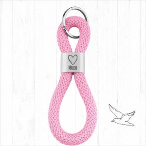 Può includere: Portachiavi in corda rosa con un dettaglio in metallo color argento. Il dettaglio presenta un cuore e il nome "Marco". Un portachiavi argentato è attaccato. Un disegno a tratto di un uccello è sullo sfondo.