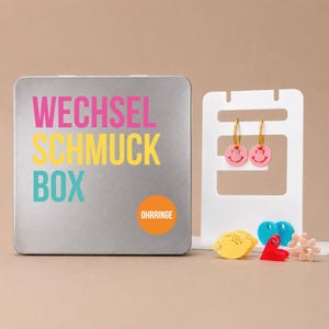 Pode incluir: Uma caixa de armazenamento de metal prateado com o texto WECHSEL SCHMUCK BOX e um círculo laranja com a etiqueta OHRRINGE. Ao lado, há um suporte branco com brincos de argola dourados com pingentes de carinha sorridente e vários pingentes.