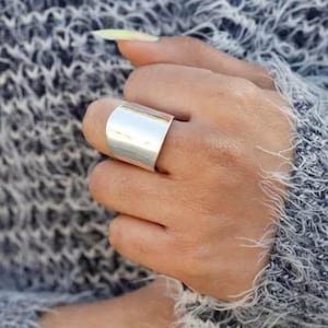 以下が含まれることがあります： 長方形のデザインの幅広のシルバーリング。 リングは指に着用され、手は質感のあるグレーと白のセーターの上に置かれています。