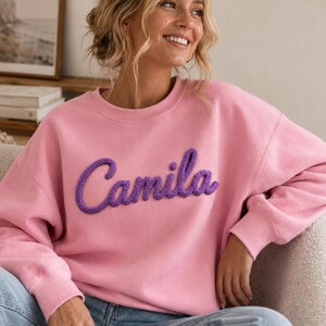 May include: A light pink long-sleeve crewneck sweatshirt featuring the name Camila written in a raised, purple, yarn-like script font across the chest. The garment has a ribbed neckline, cuffs, and hem, paired with light blue denim jeans.
