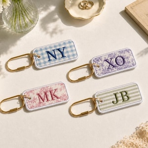 May include: Four rectangular fabric keychains with gold-toned metal rings. One features a blue gingham pattern with the letters NY, one a purple floral print with XO, one a pink floral print with MK, and one a green striped pattern with the letters JB.