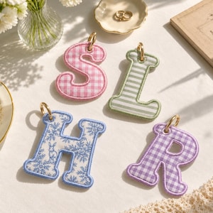 Puede incluir: Cuatro dijes de letras de tela con anillos de metal dorado. La letra S presenta un patrón de cuadros vichy rosa y blanco, la letra L tiene rayas verdes y blancas, la letra H muestra un estampado floral azul con un conejo, y la letra R es de cuadros violeta.