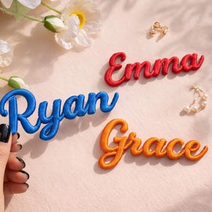 May include: Embroidered name patches in red, blue, and orange script. The names "Ryan", "Emma", and "Grace" are displayed. The patches are on a light pink surface with floral and jewelry accents.