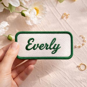 May include: A rectangular patch with the name "Everly" embroidered in green script on a white background. The patch has a green border. The image also shows floral accents and jewelry.