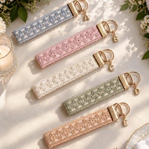 May include: Five rectangular fabric keychains featuring a floral lace pattern in blue, pink, cream, sage green, and tan. Each keychain has a gold-toned metal clasp and a small gold-toned charm shaped like the letters A, B, C, D, and E respectively.