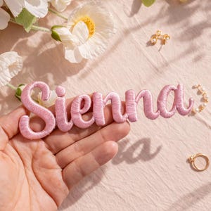 Könnte beinhalten: Ein rosa gestickter Aufnäher mit dem Namen "Sienna" in Schreibschrift. Der Aufnäher wird in einer Hand gehalten, vor einem weichen rosa Hintergrund. Schmuck und Blumen sind ebenfalls zu sehen.
