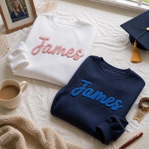 May include: A white crewneck sweatshirt with the name James embroidered in pink yarn and a navy blue crewneck sweatshirt with the name James embroidered in blue yarn, both featuring long sleeves and a soft, textured fabric construction.