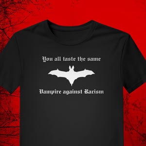 Può includere: T-shirt nera con un'immagine di pipistrello bianco e la scritta "You all taste the same" sopra il pipistrello, e "Vampire against Racism" sotto. La maglietta ha un girocollo e maniche corte.