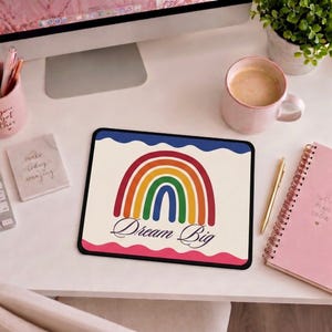 Könnte beinhalten: Rechteckiges Mauspad mit Regenbogen-Design und dem Schriftzug "Dream Big". Der Regenbogen hat rote, orange, gelbe, grüne und blaue Bögen. Das Mauspad hat einen schwarzen Rand und liegt auf einem weißen Schreibtisch.
