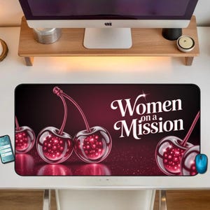 Puede incluir: Alfombrilla de escritorio rectangular con fondo rojo oscuro y la frase "Women on a Mission" en escritura blanca. La alfombrilla presenta un diseño de tres cerezas con aspecto brillante y transparente. Se ve un ratón de ordenador azul.