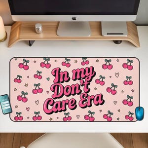 Puede incluir: Alfombrilla de escritorio rectangular rosa con un patrón de cerezas y el texto "In my Don't Care Era". La alfombrilla tiene un ratón azul, un smartphone y un personaje de dibujos animados con auriculares.