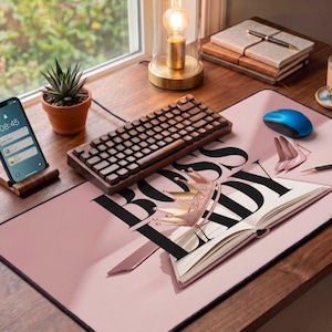 Può includere: Un'impostazione dello spazio di lavoro con un tappetino rosa con la scritta "BOSS LADY", una tastiera in legno e un mouse blu. Uno smartphone su un supporto in legno, una pianta succulenta e un bicchiere sono visibili. Una lampada e dei quaderni sono sullo sfondo.