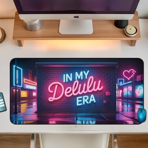 May include: A rectangular mousepad with a neon design. The design features the text "IN MY Delulu ERA" in pink and white neon, set against a backdrop of a neon-lit city street. A blue mouse and a smartphone are also visible.
